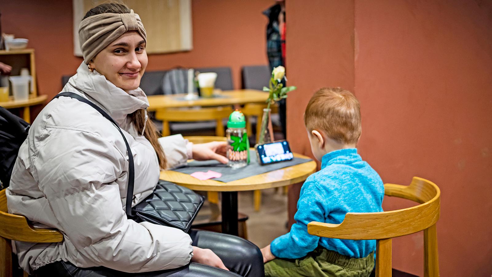 Natalia som sitter vid ett bord med sonen Matvii på Templets kår i Stockholm. Matvii sitter med ryggtavlan mot kameran och tittar på något på en mobiltelefon.