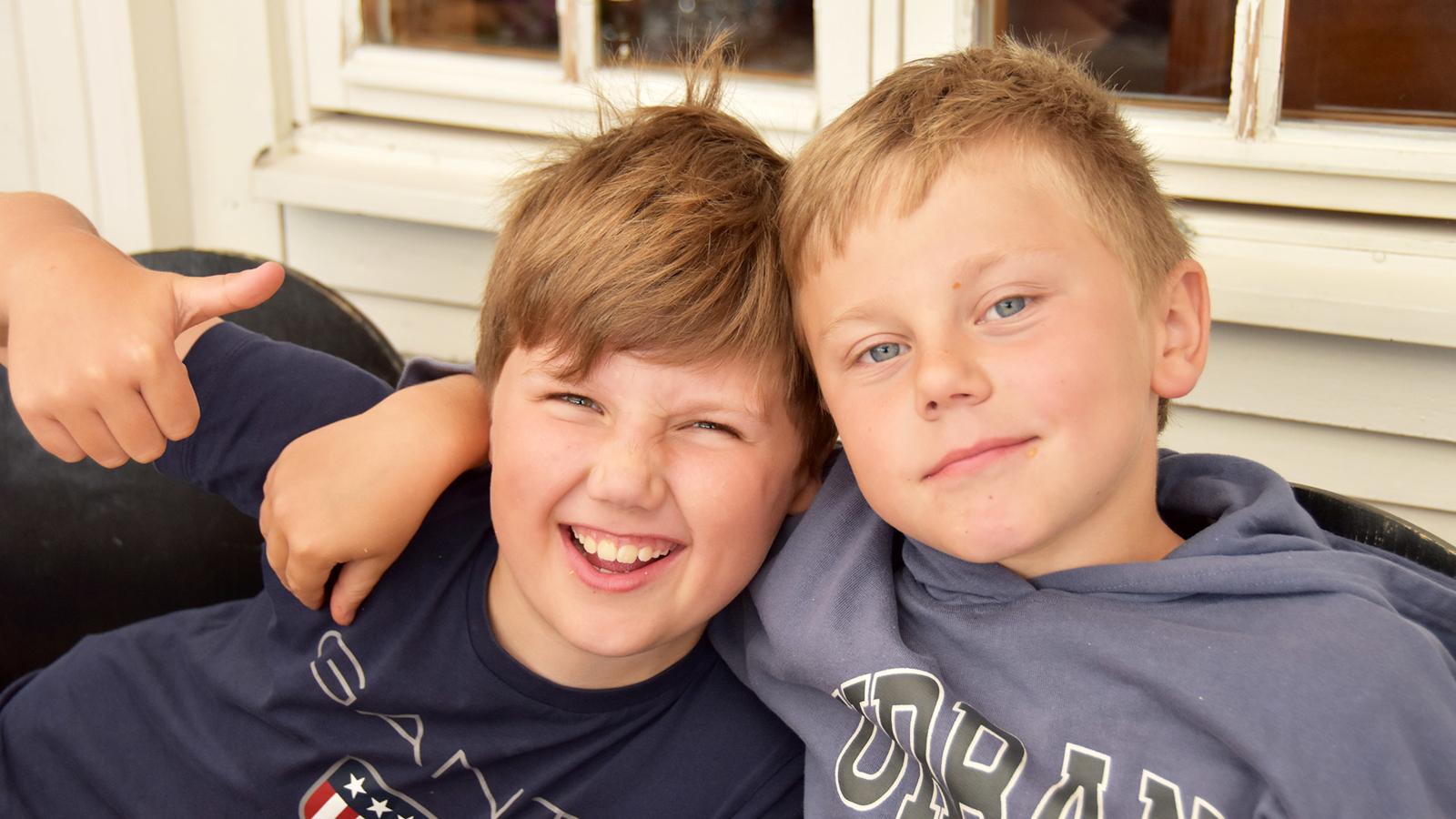 Två 10-åriga pojkar med armarna om varndras axlar tittar glatt in i kameran och gör tummen upp.