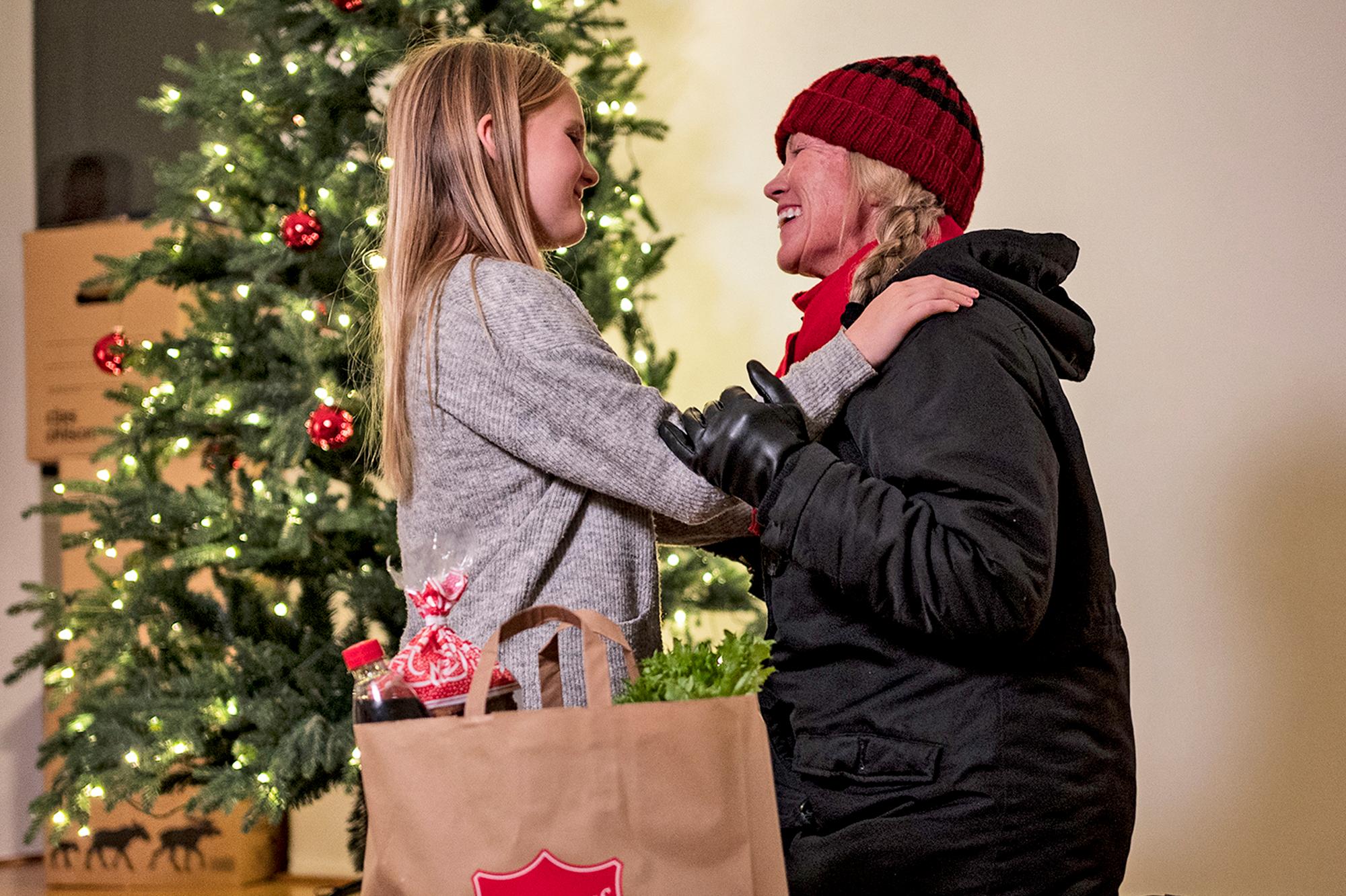 En mamma kramar om sin dotter bredvid en julgran, en kasse mat står vid deras sida. 