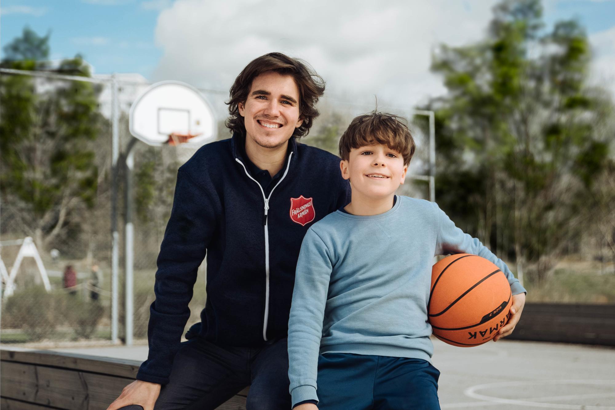 En yngre man med Frälsningsarméns sköld på jackan sitter på en bänk vid en basketplan med en liten pojke som håller i en basketboll. Båda ler in i kameran.