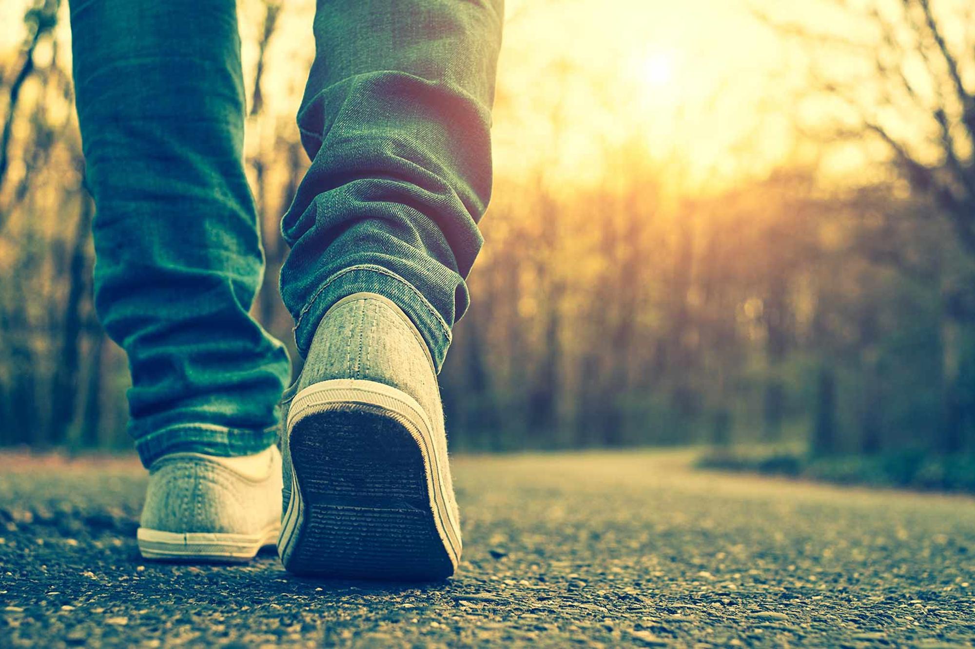 Fokus på skorna på en person som går på en väg med en solnedgång i bakgrunden.