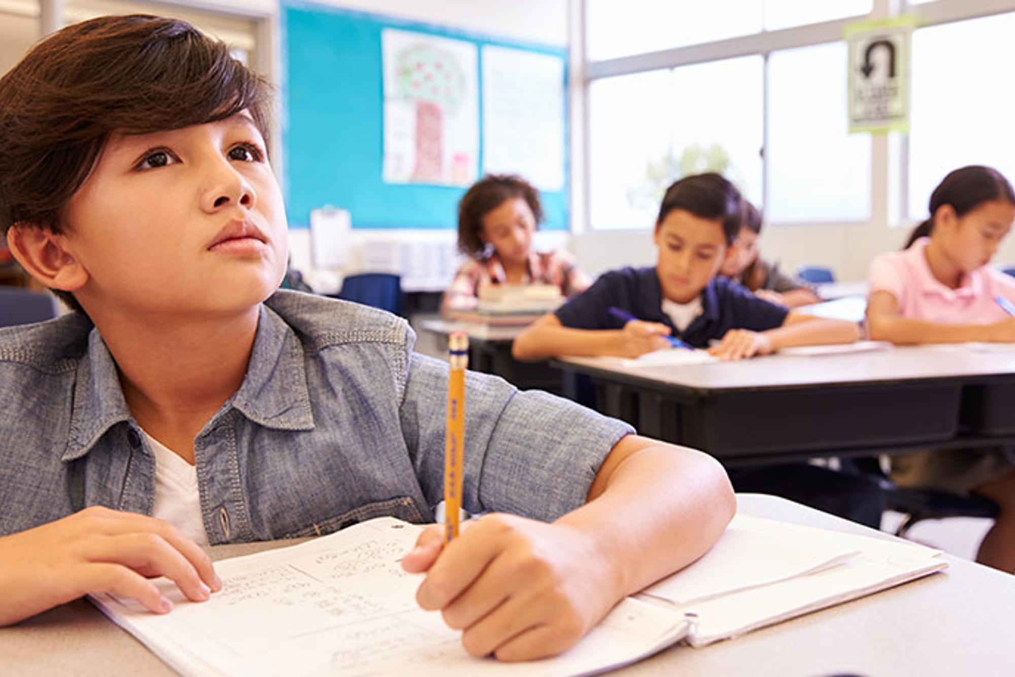 En pojke som sitter i ett klassrum och skriver i en skolbok. I bakgrunden syns andra elever som också antecknar.