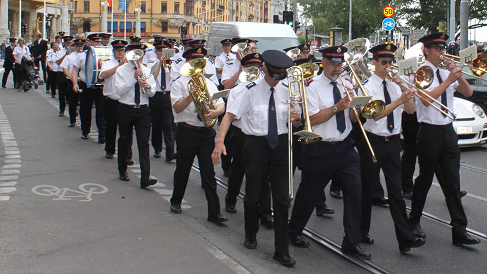 Musikkåren marscherar på Stockholms gator. De spelar på olika typer av instrument.