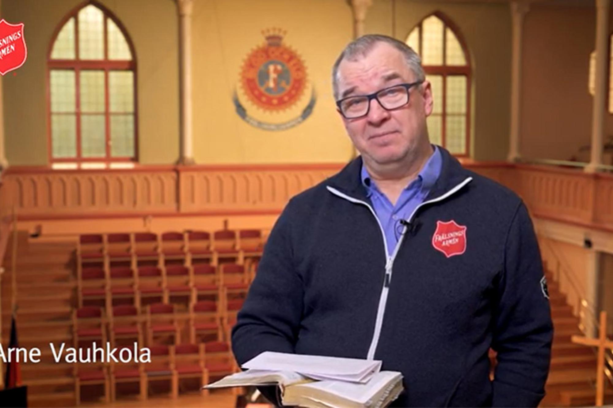 Arne Vauhkola står i en kyrka med en uppslagen bok i ena handen och blicken in i kameran.