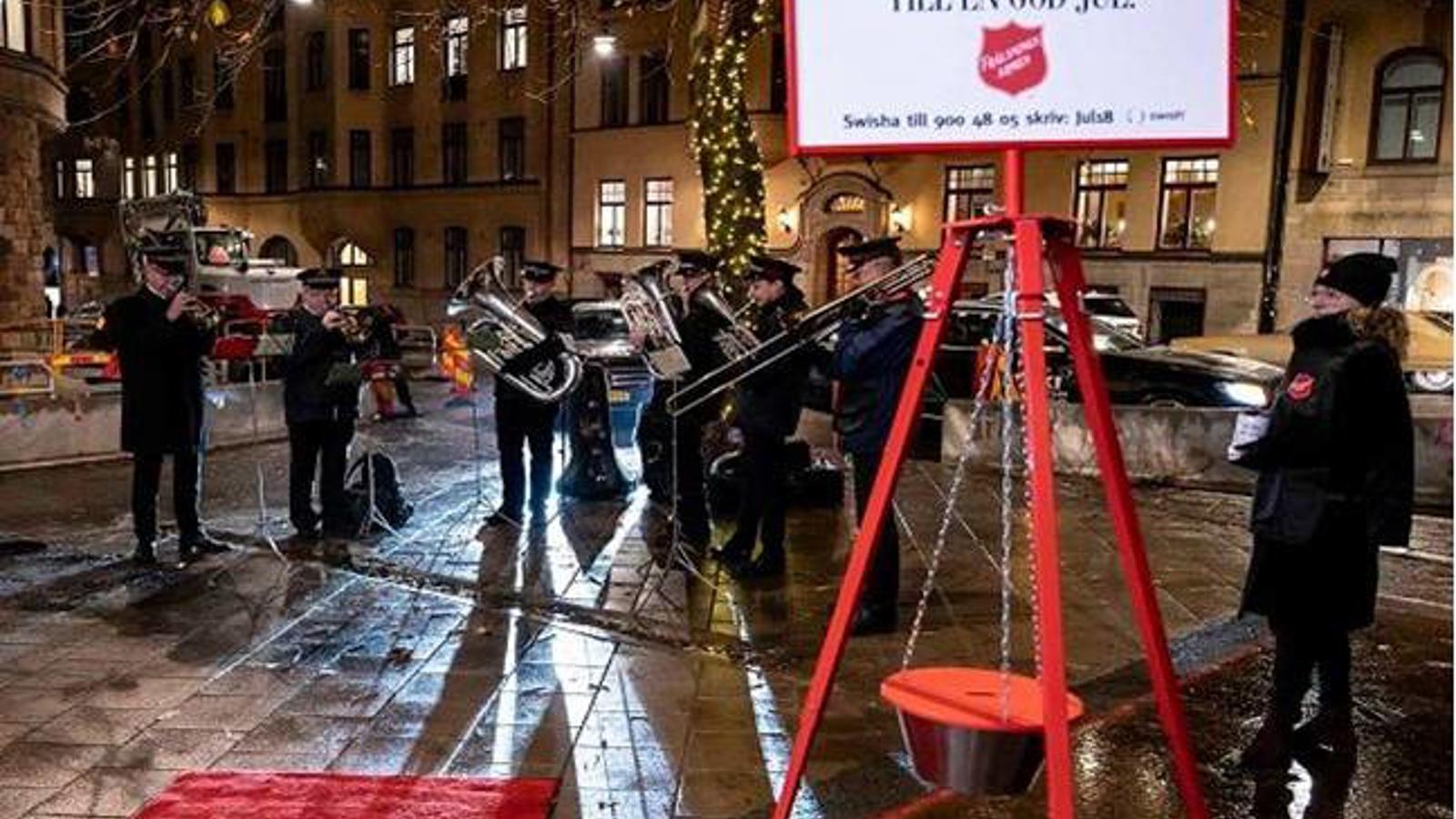 En Frälsningsarméns-skylt med texten "Ge fler möjlighet till en God Jul". Bakom skylten står personer från Frälsningsarmén och spelar instrument.