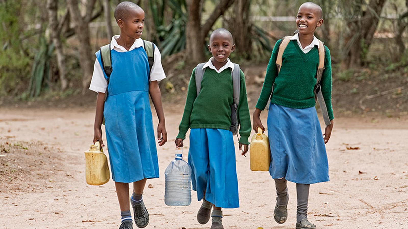 Tre glada flickor i Kenya går tillsammans med ryggsäck på ryggen med varsin plastdunk i handen som de ska fylla med vatten.