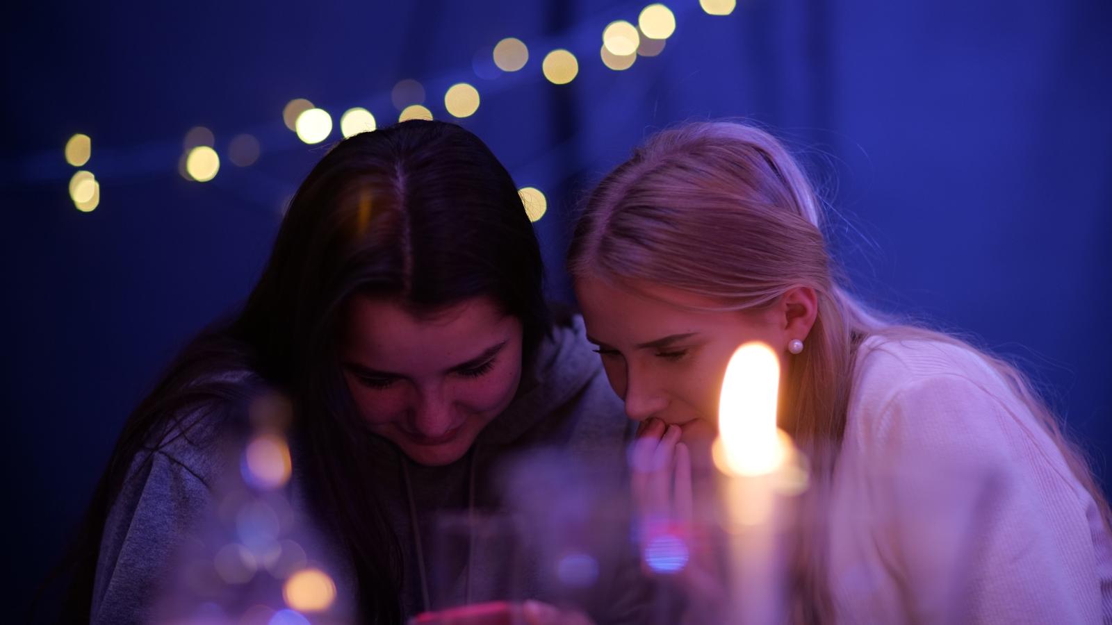 Två unga kvinnor som sitter och kollar på en mobiltelefon. På bordet framför dem står det ett tänt ljus.
