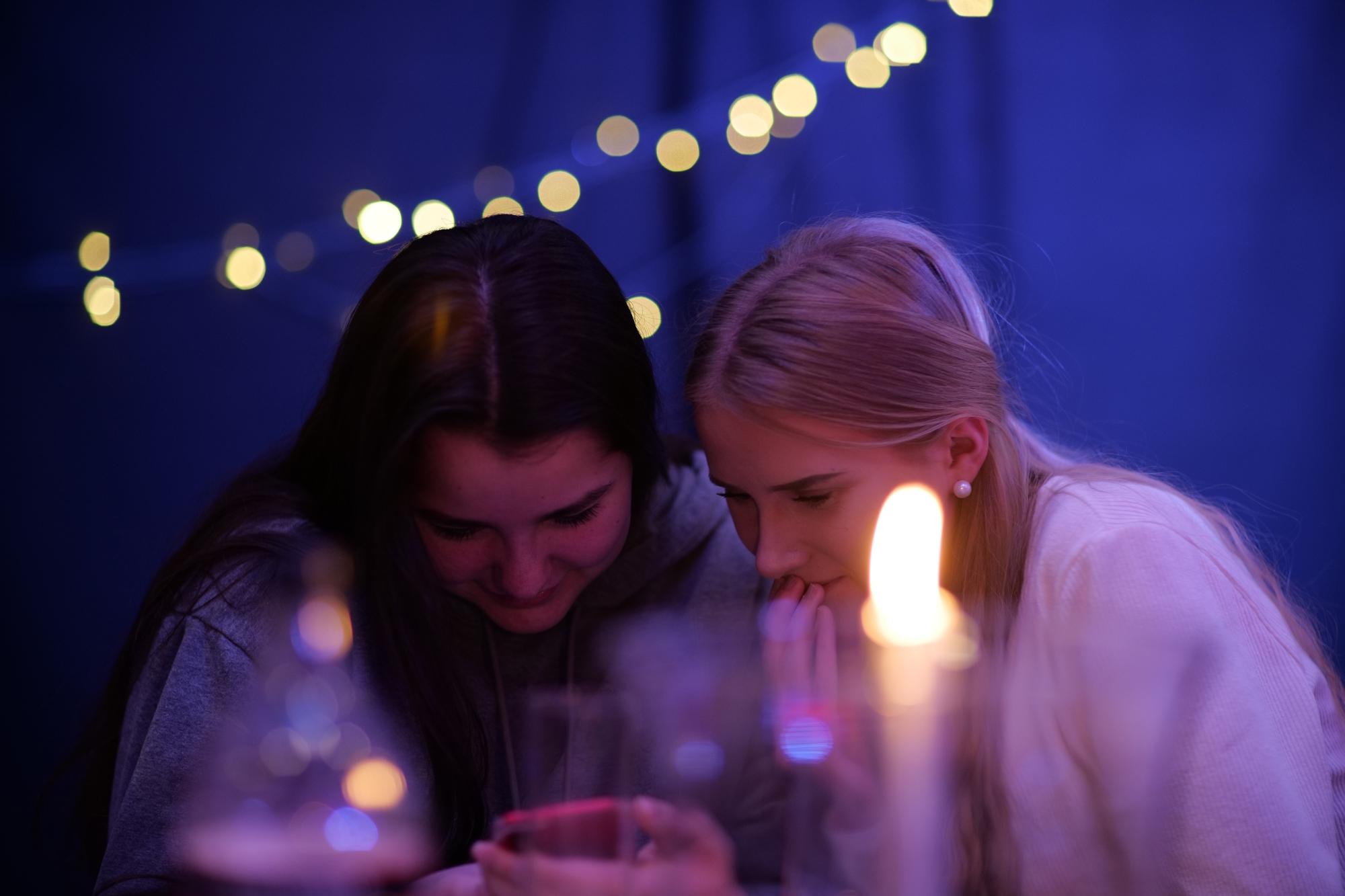 Två unga kvinnor som sitter och kollar på en mobiltelefon. På bordet framför dem står det ett tänt ljus.