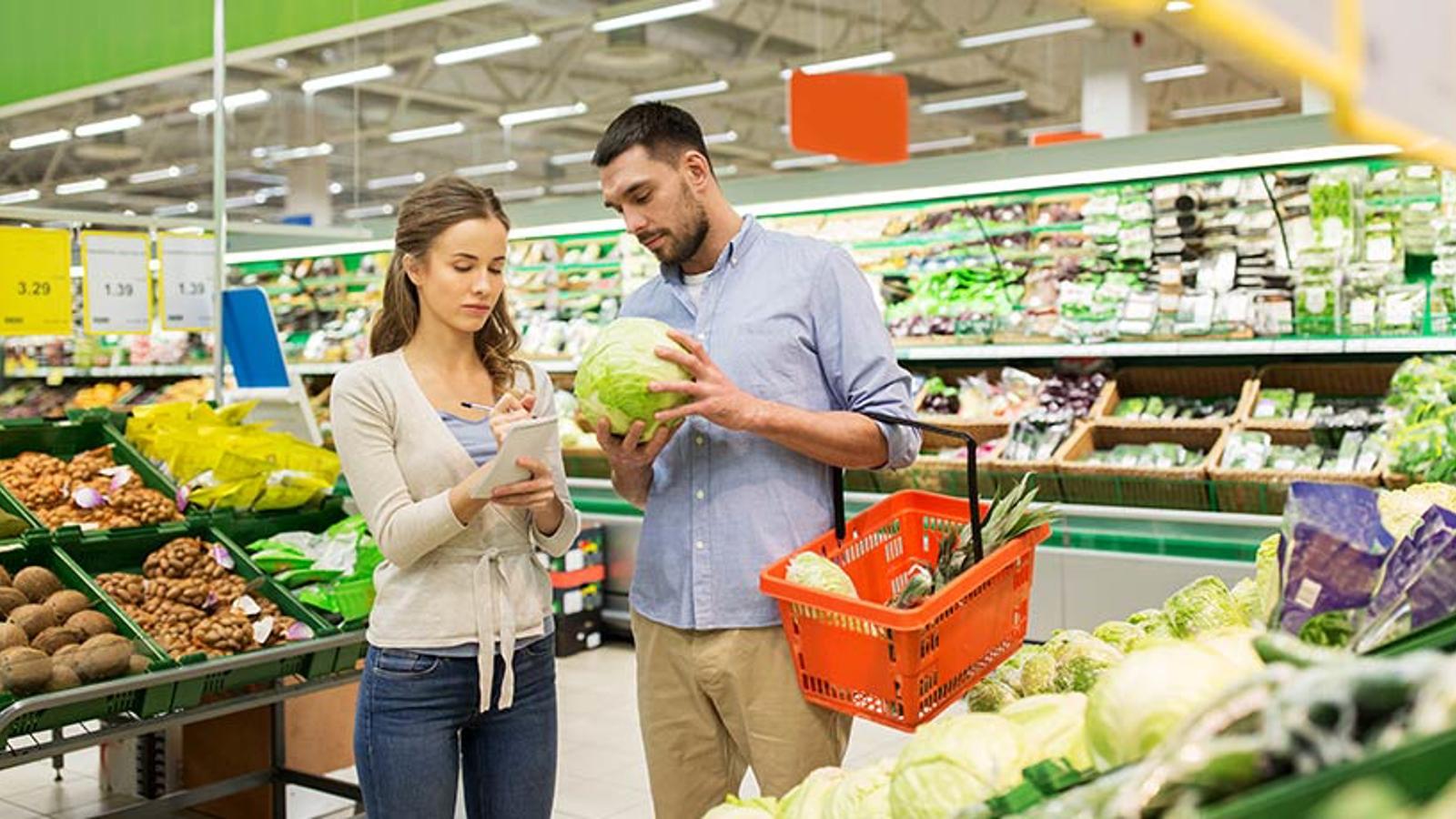 En man och en kvinna som står och kollar på grönsaker i en matbutik. Kvinnan står och bockar av en inköpslista.