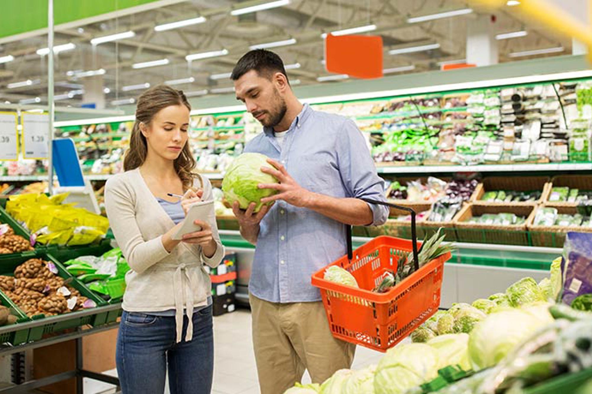 En man och en kvinna som står och kollar på grönsaker i en matbutik. Kvinnan står och bockar av en inköpslista.