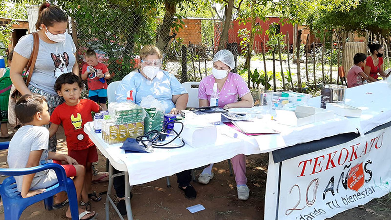 Tre kvinnor med munskydd och två pojkar som sitter vid ett långt bord. På bordet hänger det ett skynke med texten "Teko katu" som betyder ”ett fullständigt liv” på Paraguays lokala språk, guaraní.