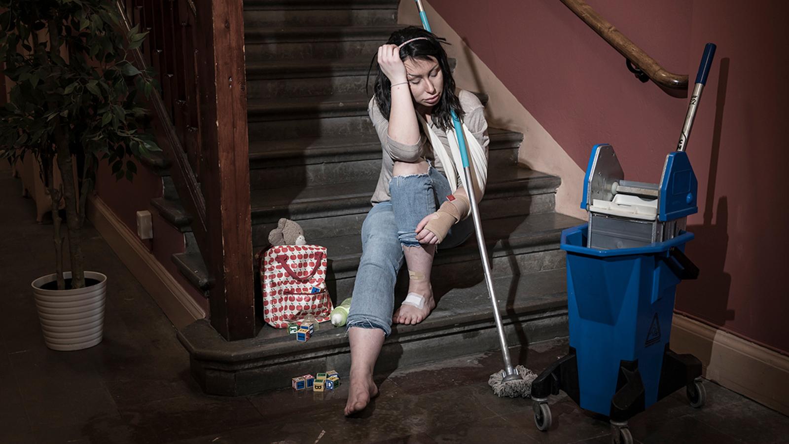 En ung och utmattad kvinna som sitter i en trappuppgång. Hon har ett plåster på ena foten och hennes ena handled är inlindad i bandage. Runtom henne står det städutrutsning.