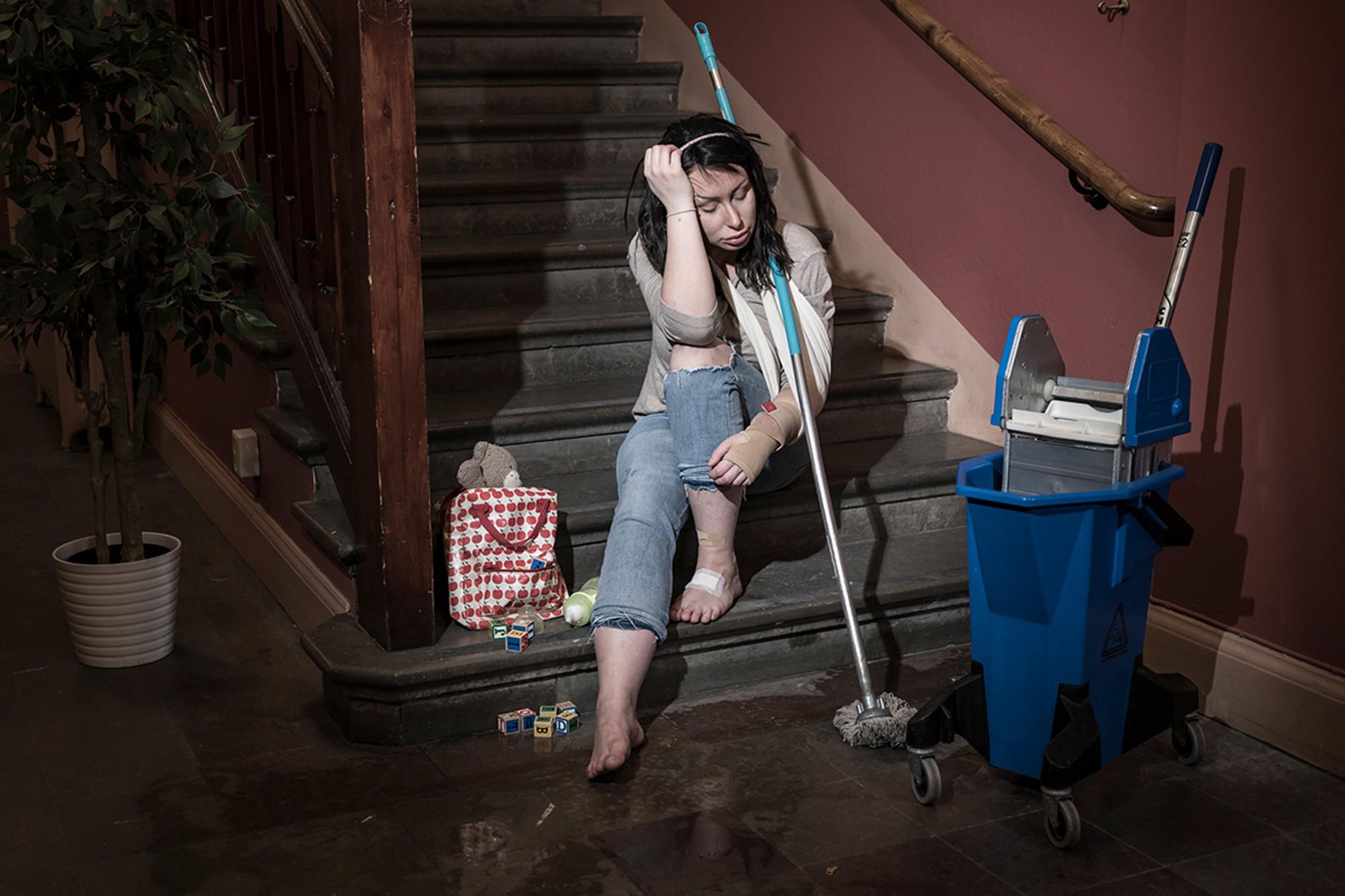 En ung och utmattad kvinna som sitter i en trappuppgång. Hon har ett plåster på ena foten och hennes ena handled är inlindad i bandage. Runtom henne står det städutrutsning.