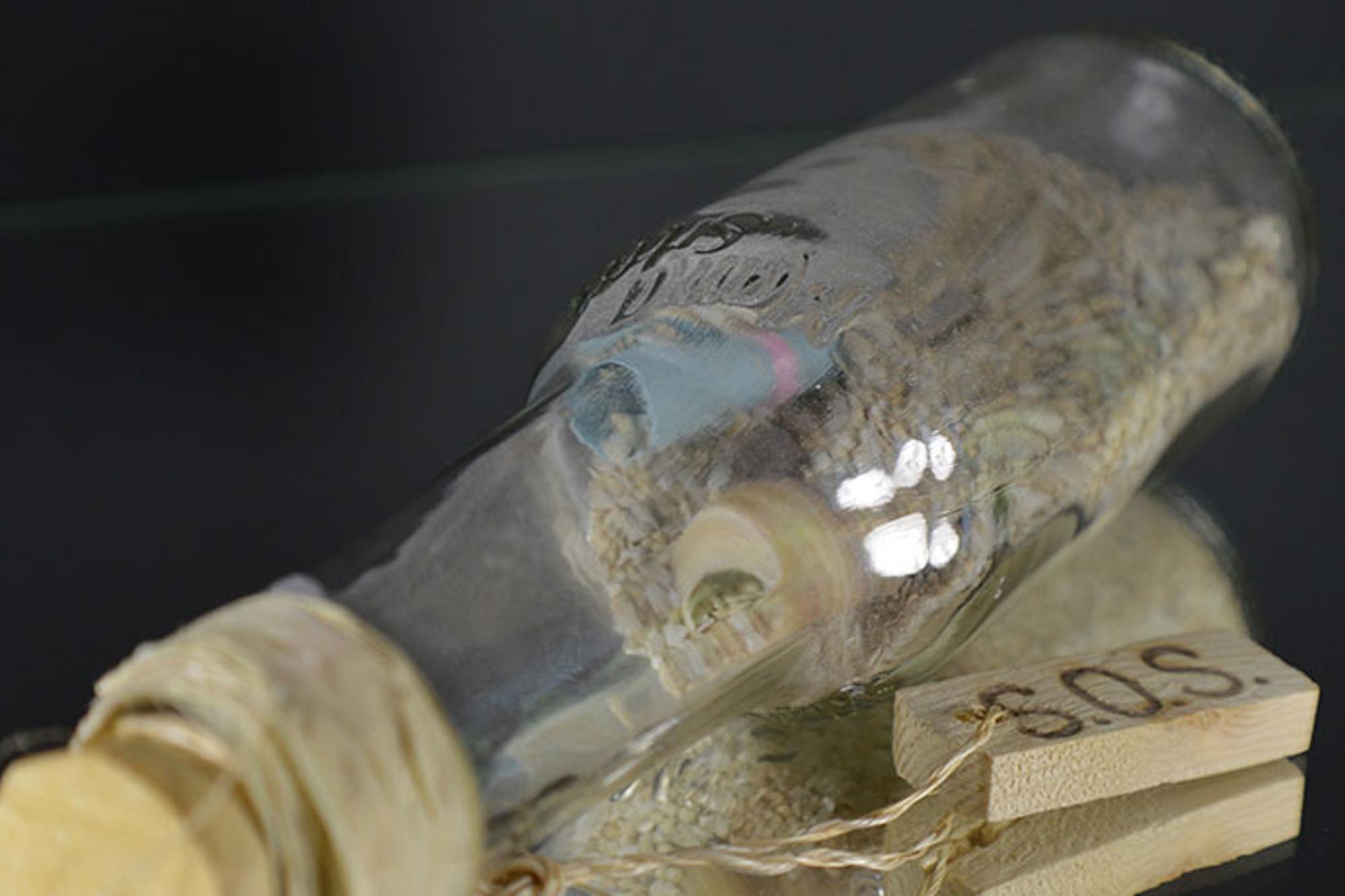 En glasflaska med en lapp i. På flaskan hänger det ett snöre som sitter fast i en liten träbit med texten "SOS".