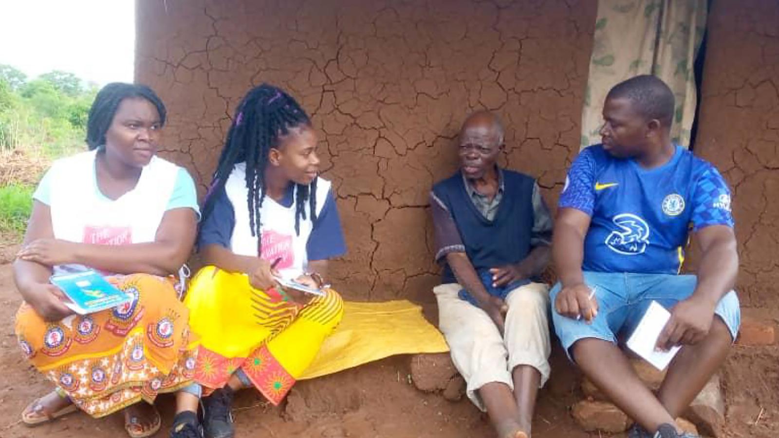 Personal från Frälsningsarmén i Malawi sitter vid en lerfärgad vägg med sprickor i tillsammans med en äldre man och samtalar.