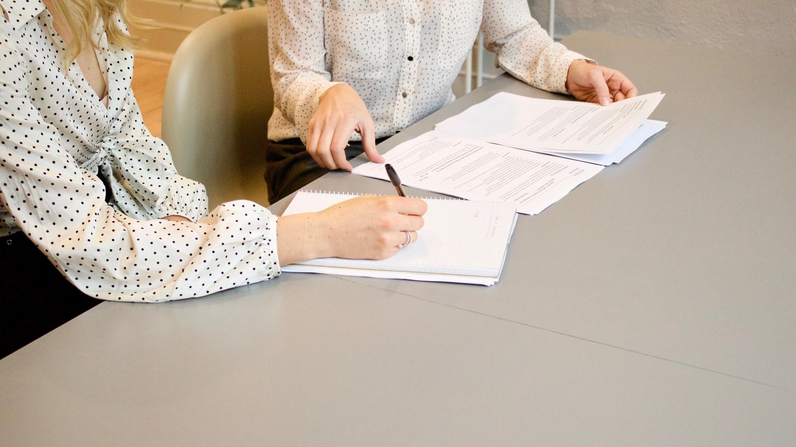 Två personer som sitter vid ett bord, den ena har papper framför sig och den andra skriver i ett anteckningsblock.