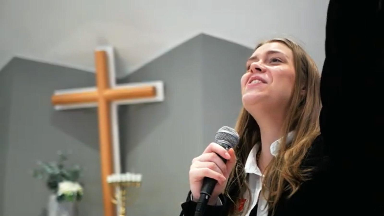 Ung kvinna sjunger med mikrofon i ena handen och andra handen lyft ovanför huvudet. Ett kors står i bakgrunden.