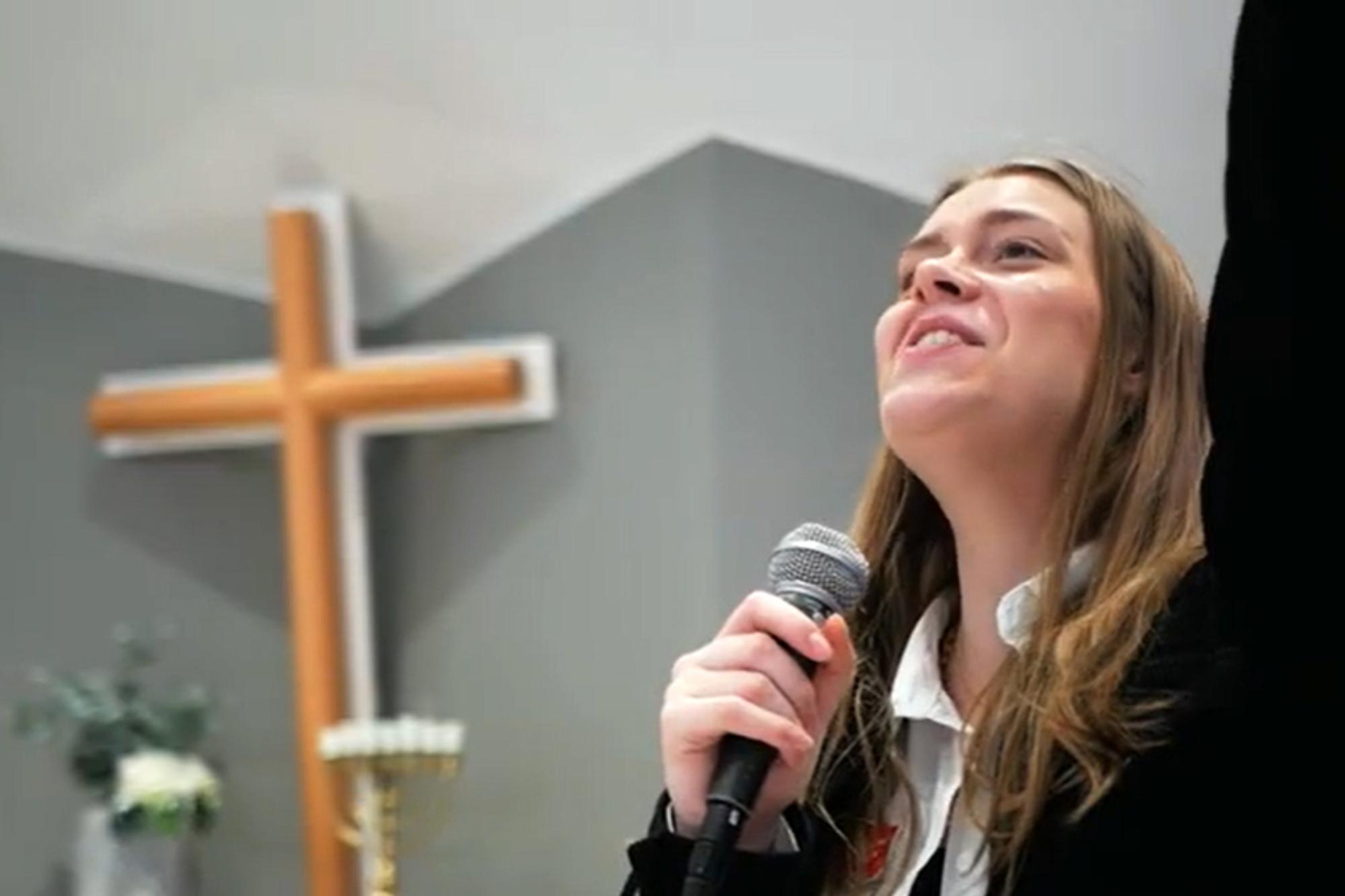 Ung kvinna sjunger med mikrofon i ena handen och andra handen lyft ovanför huvudet. Ett kors står i bakgrunden.