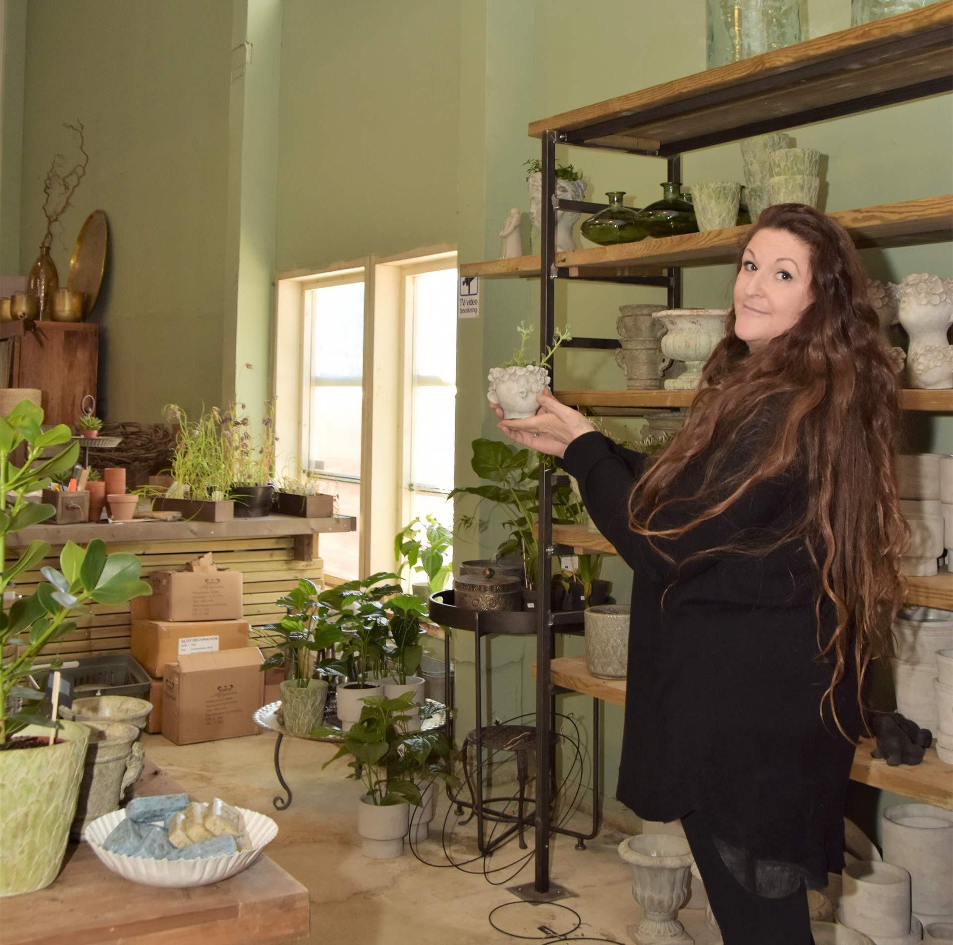 Martina håller upp en kruka med en liten växt och tittar in i kameran. I bakgrunden syns olika krukor, dekorationer och växter mot en grön vägg.