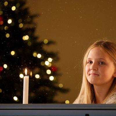 En flicka står i fönstret bredvid ett tänt ljus. En julgran syns i bakgrunden.