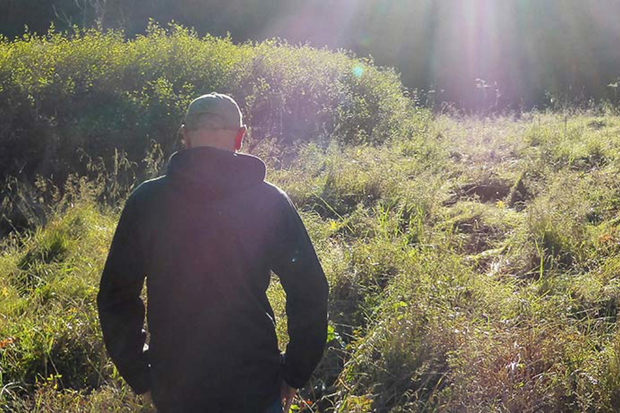En man med ryggtavlan mot kameran. Han står ute i naturen och i bakgrunden syns solstrålar.