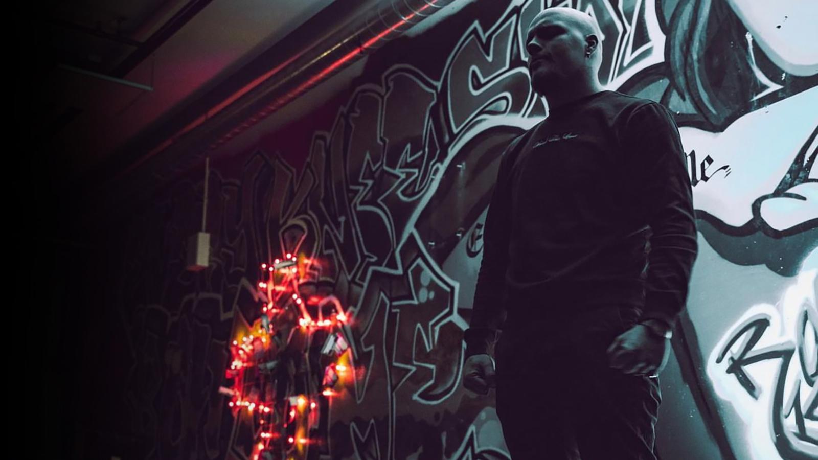 Simon Nygren står i ett mörkt rum med knutna händer framför en vägg med graffiti, bredvid honom står ett kors klätt med en ljusslinga.