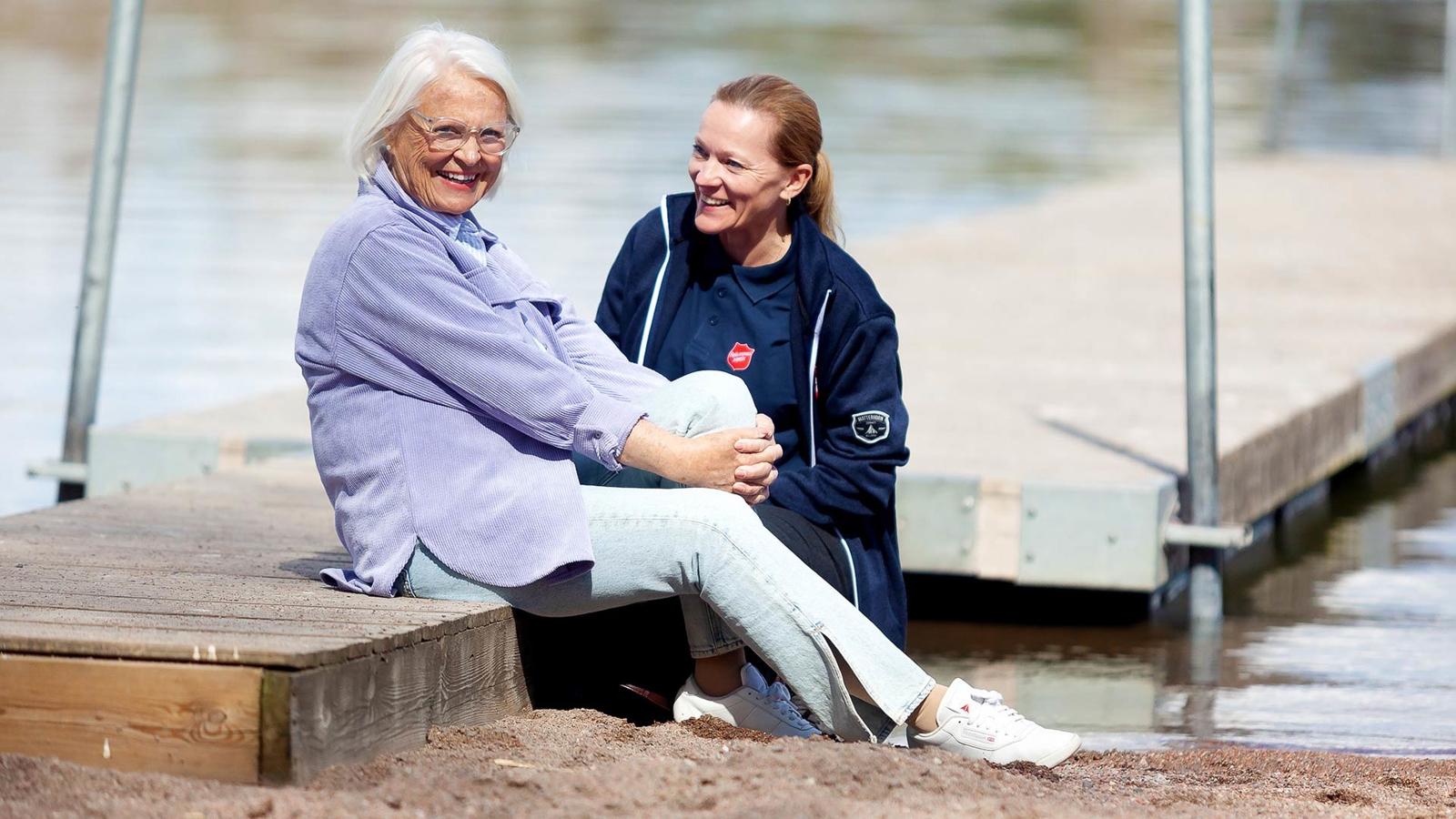 En äldre kvinna sitter leende på en brygga  vid en sjö tillsammans med en yngre kvinna som har Frälsningsarméns sköld på tröjan.