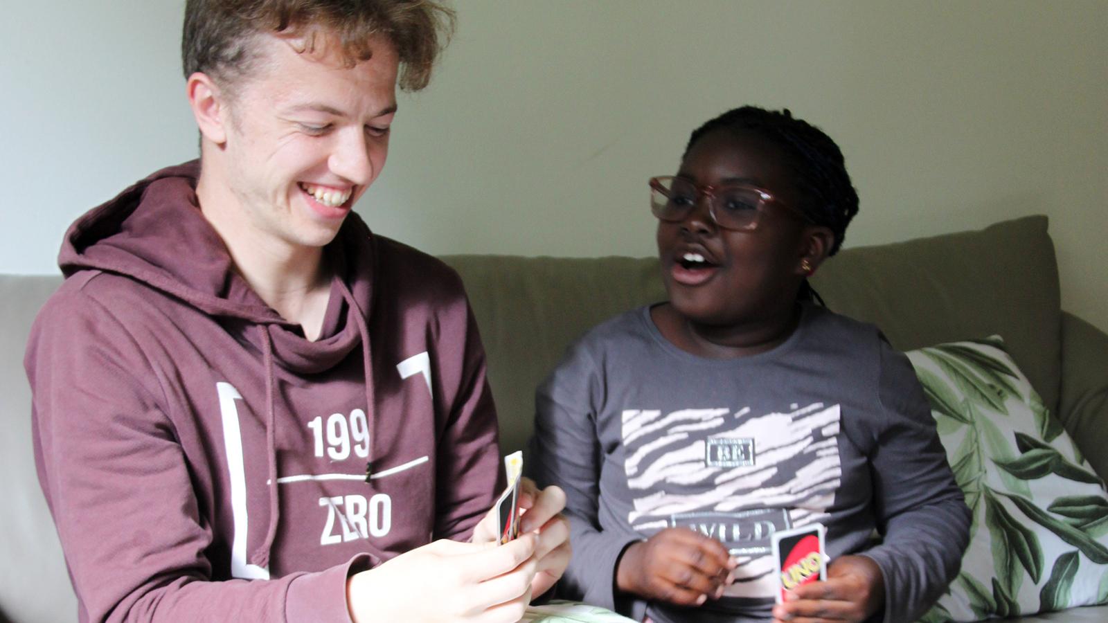 En ung kille och en liten flicka sitter i en soffa och spelar kortspelet UNO - de skrattar och ser glada ut. 