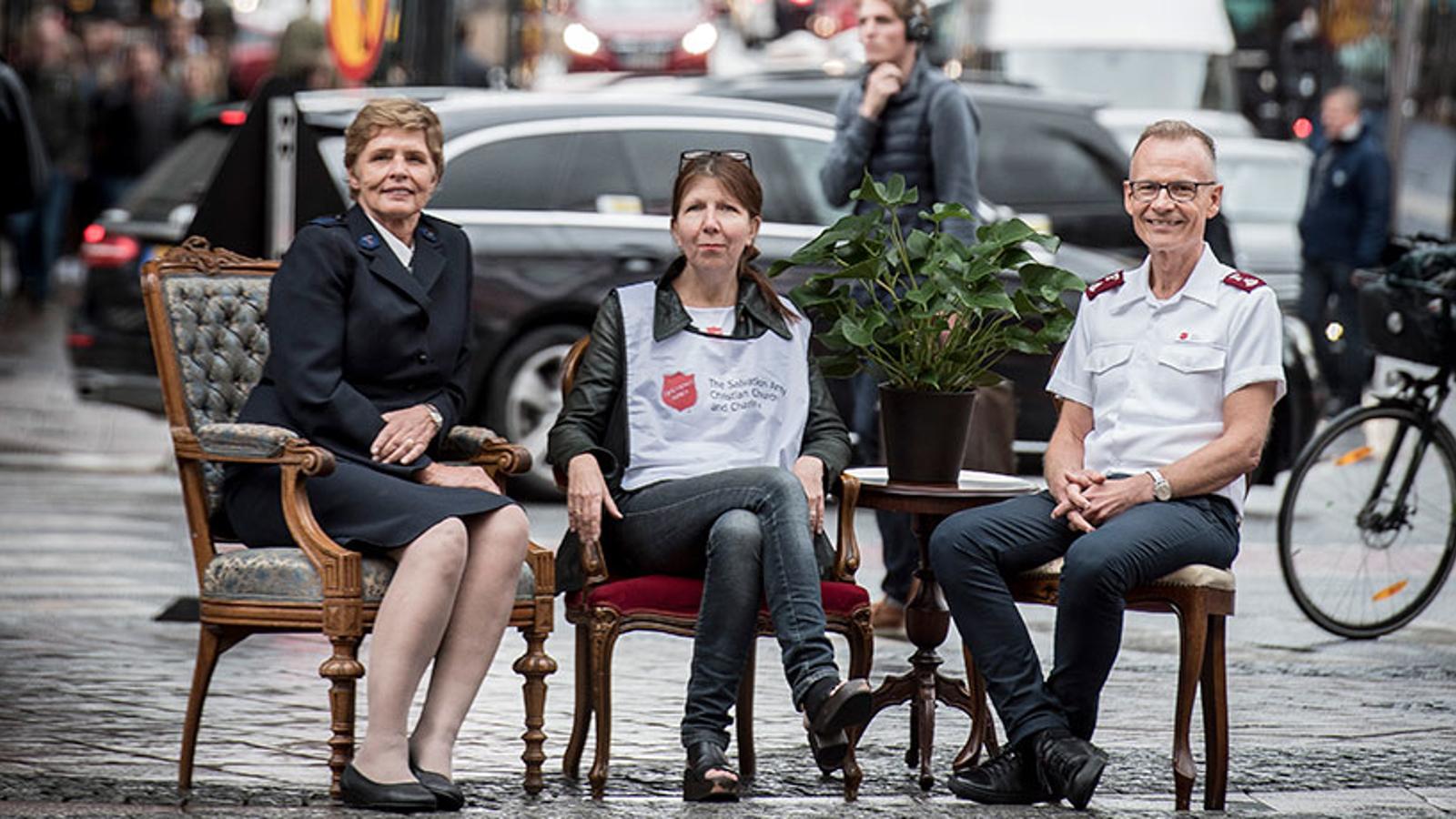 Två kvinnor och en man i Frälsningsarméns uniform resp profilkläder sitter i varsin stol ute på ett torg i stadsmiljö. Bilar ,fotgängare och cyklister i bakgrunden.