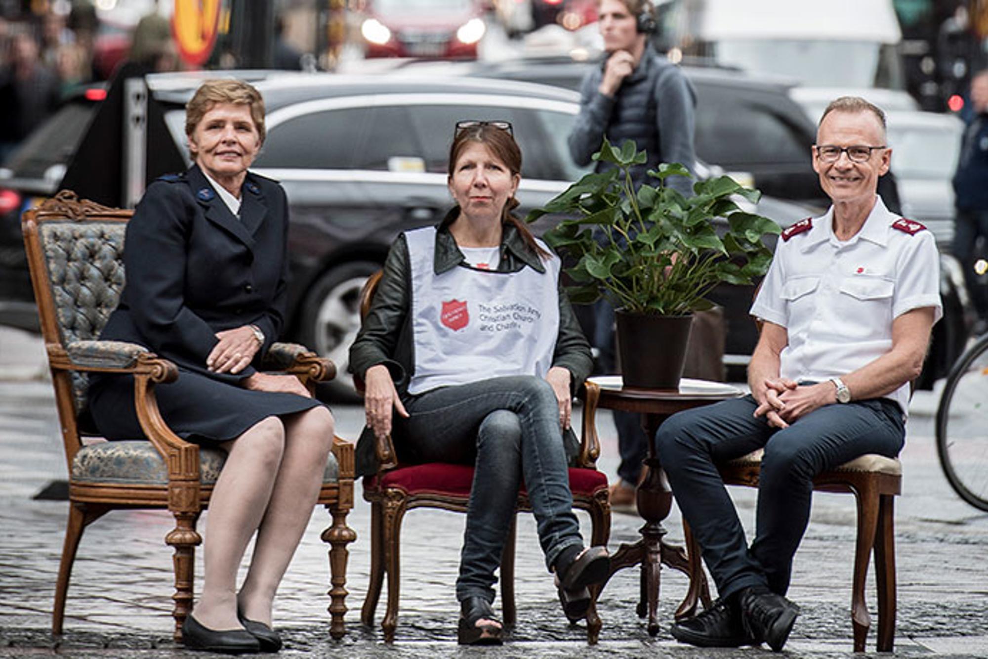 Två kvinnor och en man i Frälsningsarméns uniform resp profilkläder sitter i varsin stol ute på ett torg i stadsmiljö. Bilar ,fotgängare och cyklister i bakgrunden.