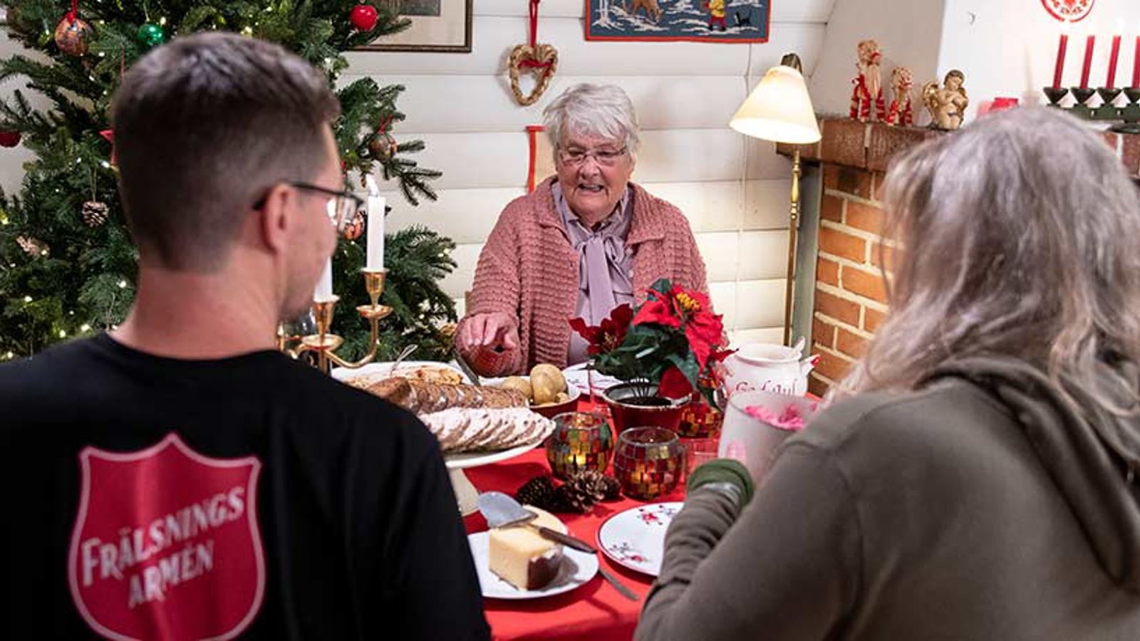 Ryggtavlan på en man från Frälsningsarmén som sitter vid ett julbord och äter julmat tillsammans med två äldre damer. I bakgrunden står det en pyntad julgran. 
