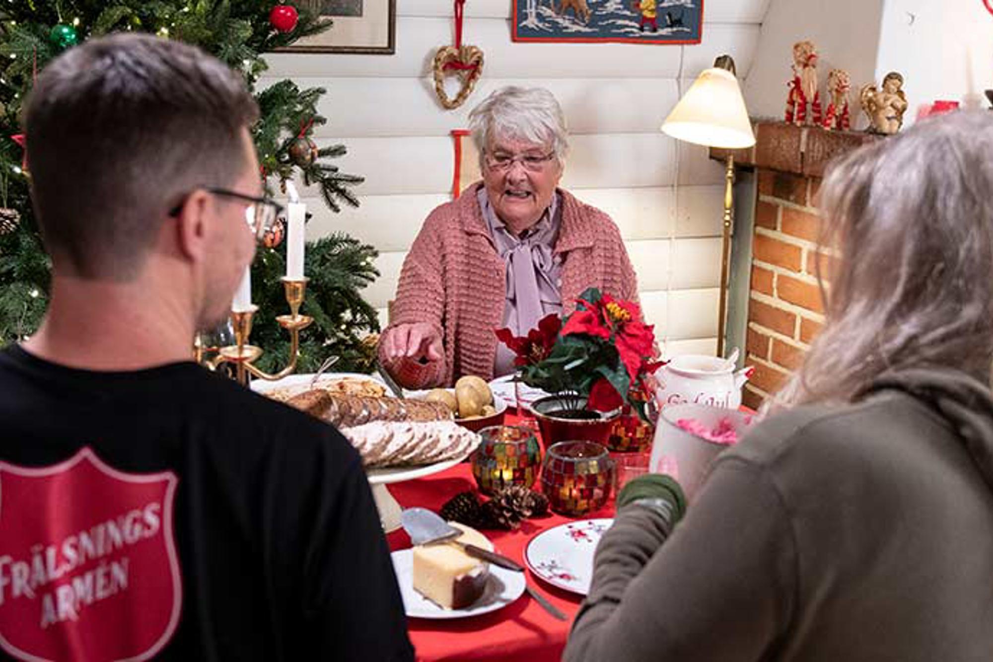 Ryggtavlan på en man från Frälsningsarmén som sitter vid ett julbord och äter julmat tillsammans med två äldre damer. I bakgrunden står det en pyntad julgran. 