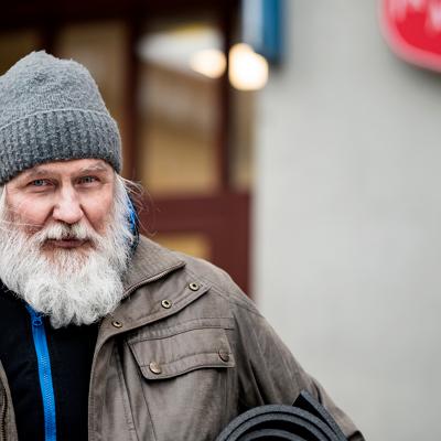 En vinterklädd äldre man med skägg står på gatan utanför en dörr med Frälsningsarméns sköld. Han har ett liggunderlag under armen.