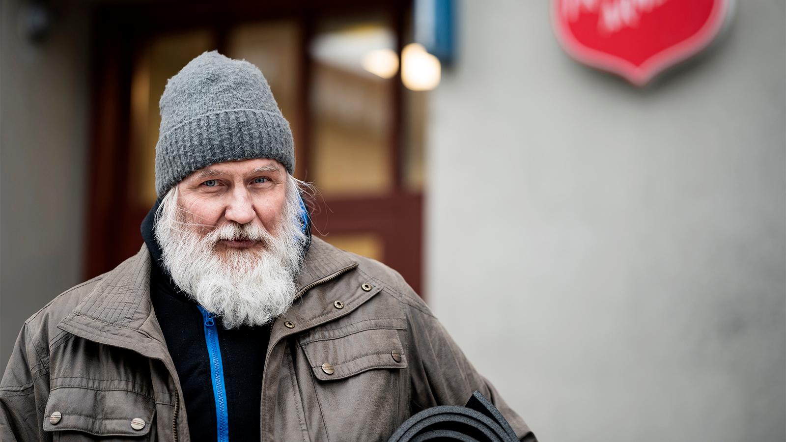 En vinterklädd äldre man med skägg står på gatan utanför en dörr med Frälsningsarméns sköld. Han har ett liggunderlag under armen.