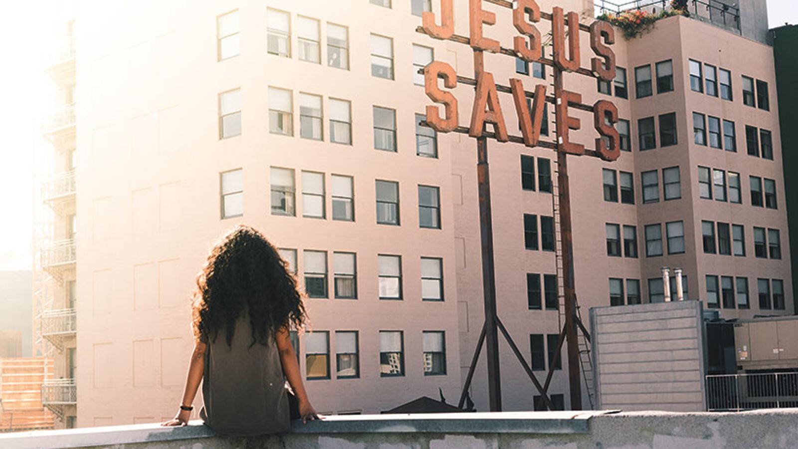 Ryggtavlan på en kvinna som sitter på kanten av en stenmur med blicken mot en byggnad med texten "Jesus Saves". 