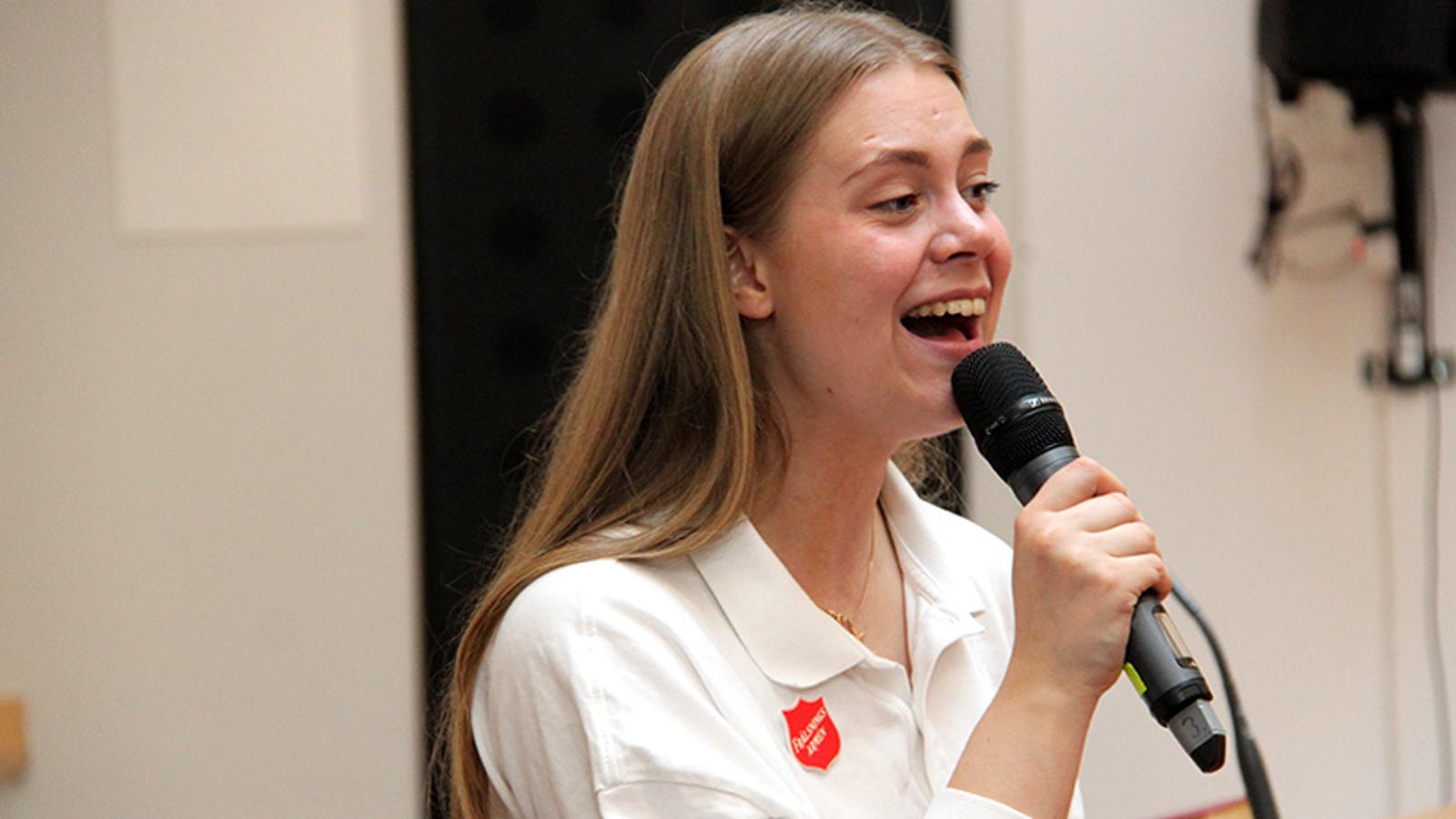 En ung kvinna med Frälsningsarmén sköld på tröjan som sjunger i en mikrofon.