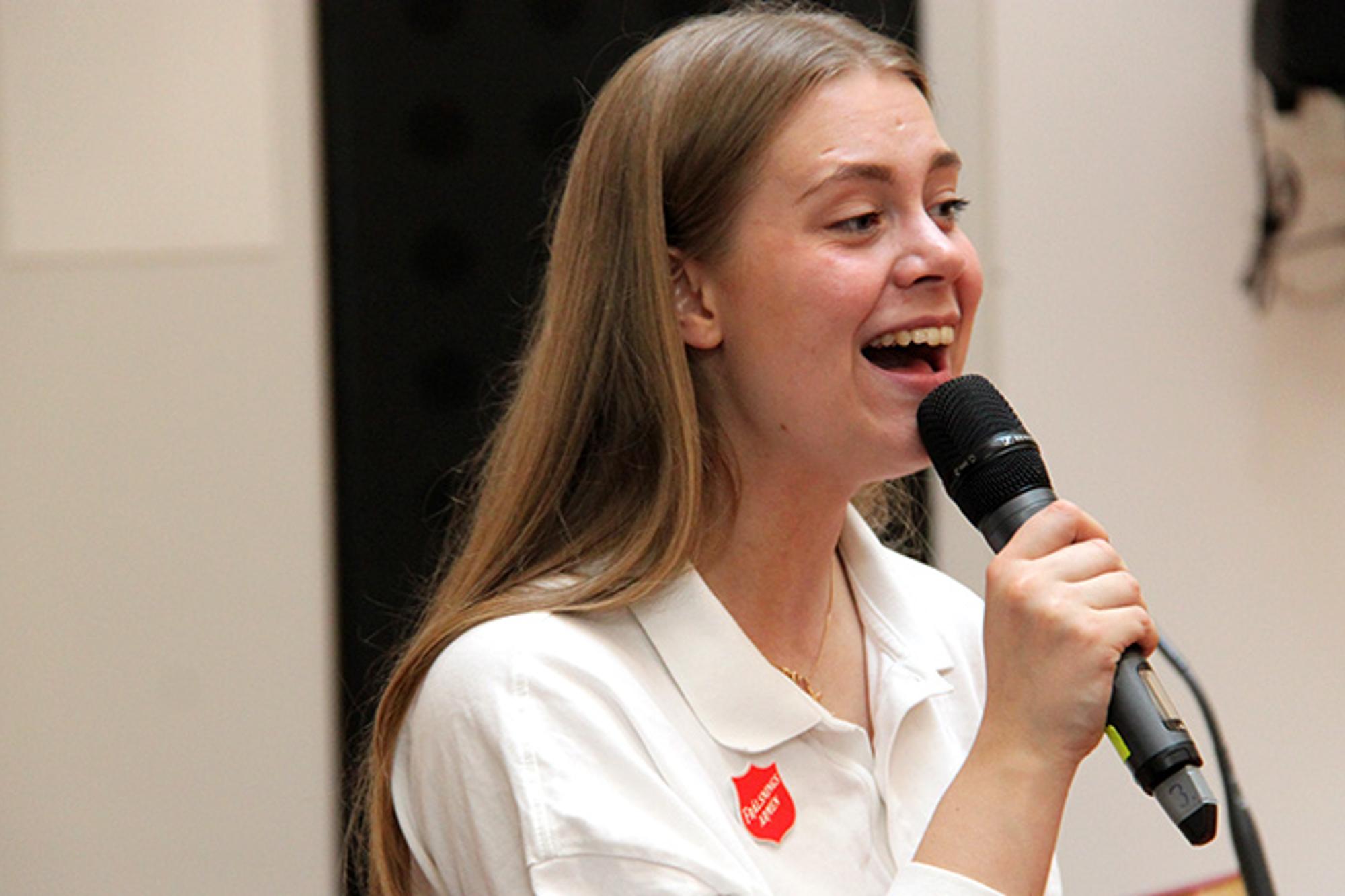 En ung kvinna med Frälsningsarmén sköld på tröjan som sjunger i en mikrofon.