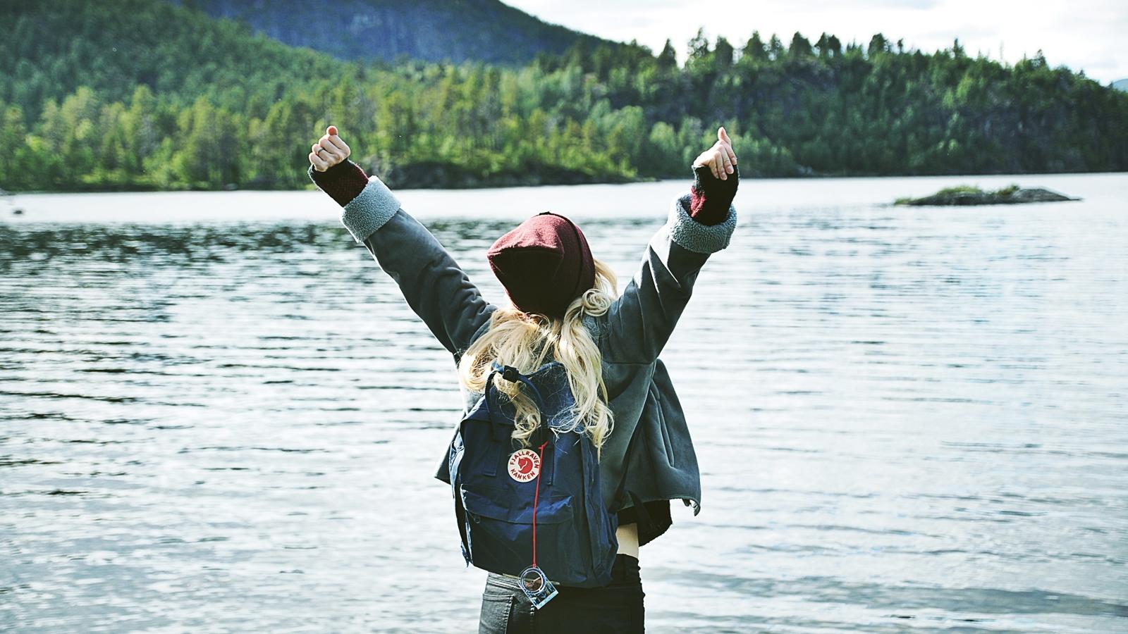 Ryggtavlan på en kvinna med ryggsäck som står vänd mot en sjö och sträcker armarna upp i luften.