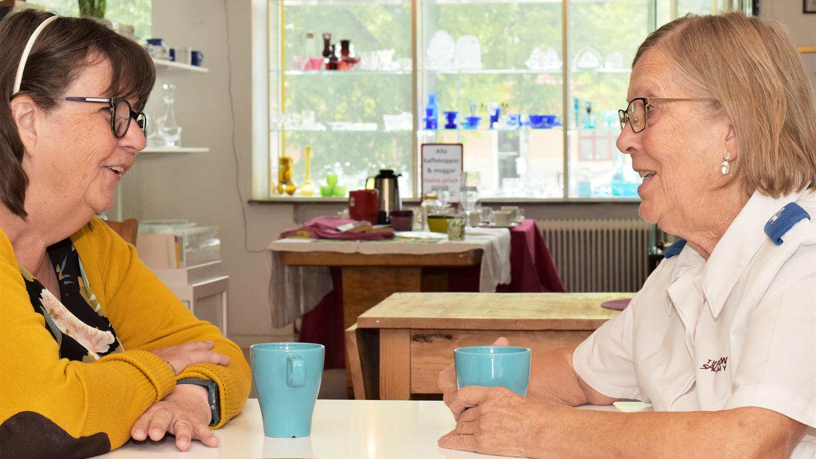Elisabeth och Solveig sitter mitt emot varandra och samtalar vid ett kafébord med kaffekoppar i händerna.