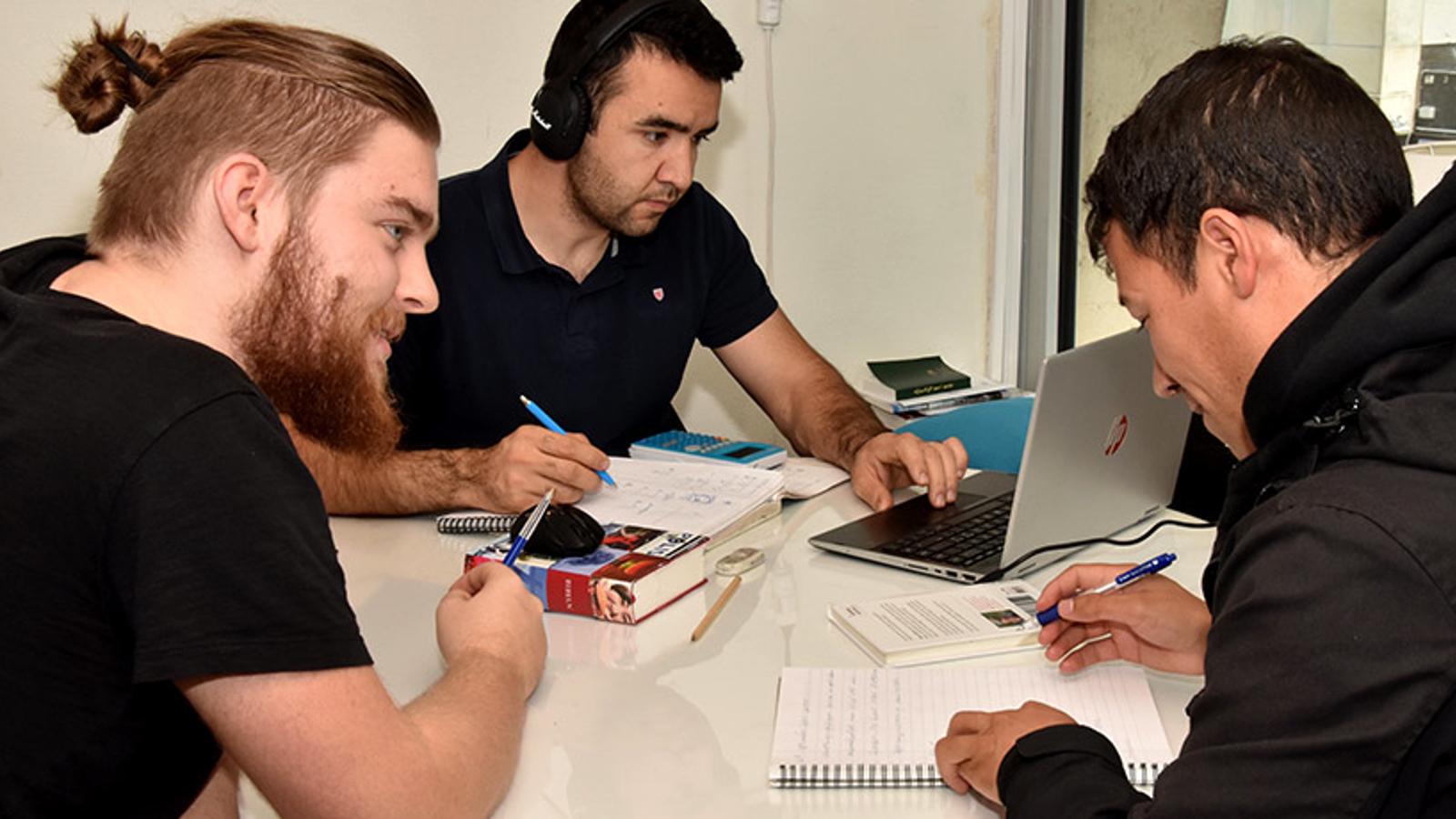Tre unga män runt ett bord som studerar körkortsteori. De sitter med en dator, anteckningsblock och pennor.