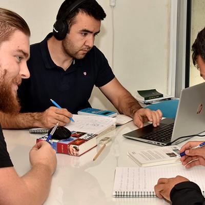 Tre unga män runt ett bord som studerar körkortsteori. De sitter med en dator, anteckningsblock och pennor.
