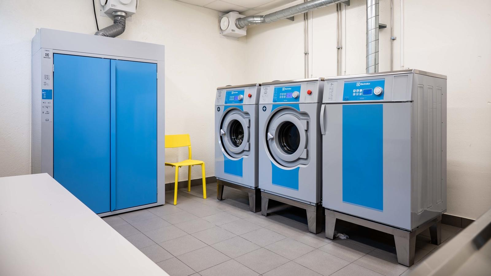 Tvättstuga, med två tvättmaskiner, en torktumlare och ett torkskåp, alla är ljusblå. Mellan tvättmaskinerna och torkskåpet står en klargul stol. 