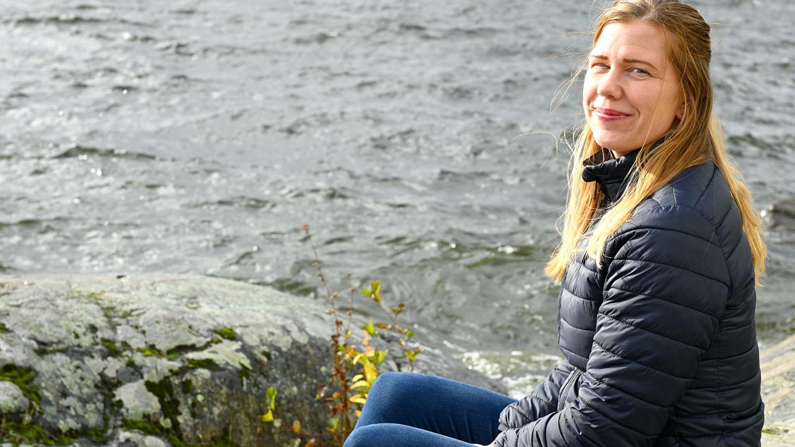 Helen sitter på klipporna vid en sjö och tittar in i kameran.