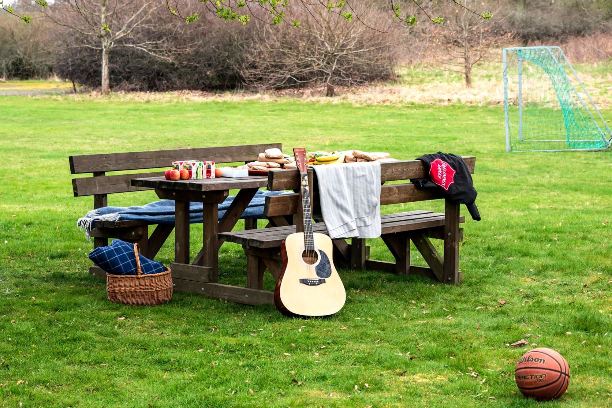 Ett picknickbord med smörgåsar, festis, filtar, en gitarr, en boll och ett fotbollsmål i bakgrunden. En jacka med Frälsningsarméns sköld ligger på bänken