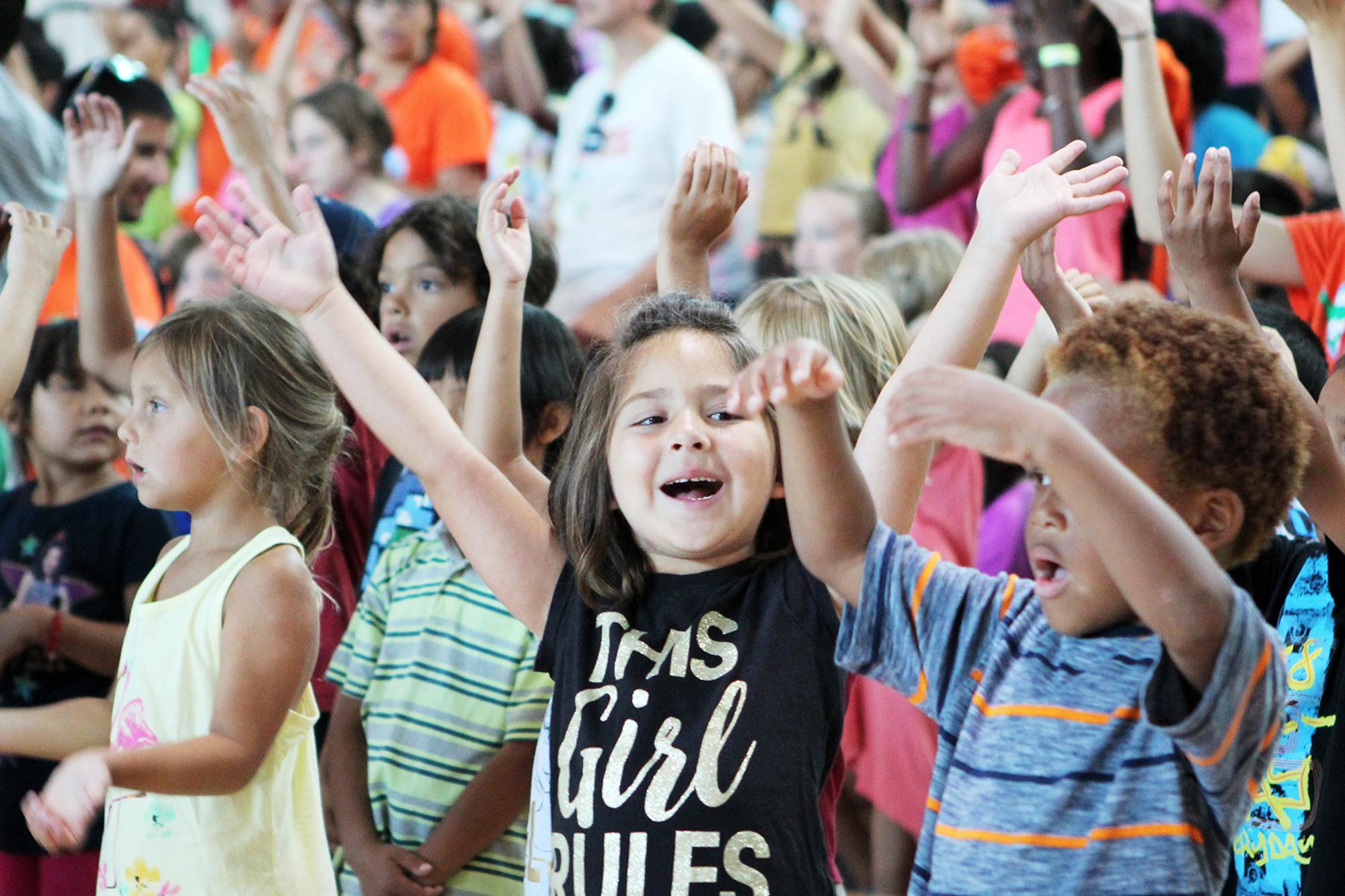 Många glada barn står i någon typ av arena med händerna i luften.