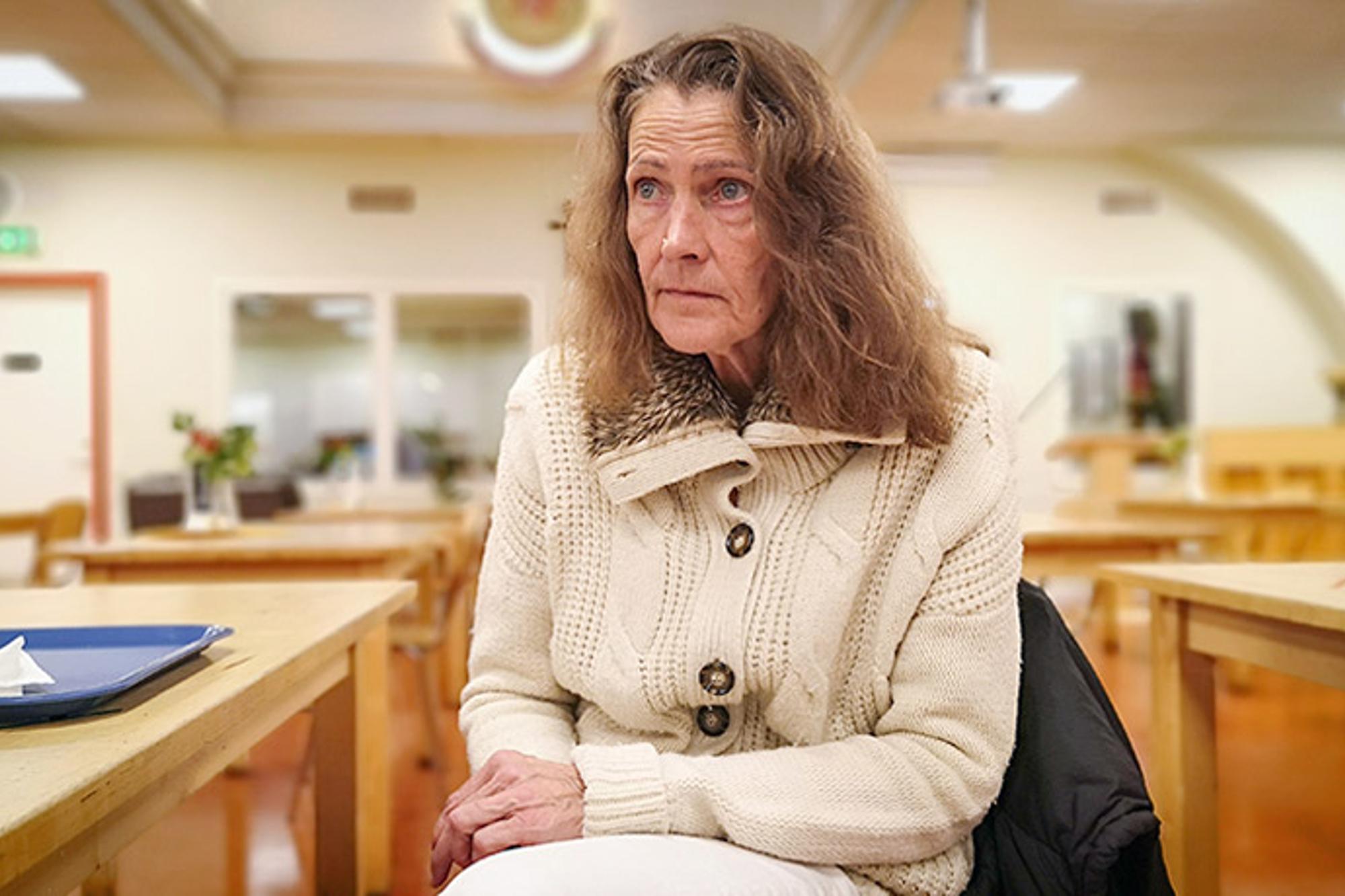 En kvinna, Sussie, som tittar till vänster om kameran. Hon sitter vid ett bord i sociala centrets lokal. På bordet står det en blå bricka och i bakgrunden syns flera bord.