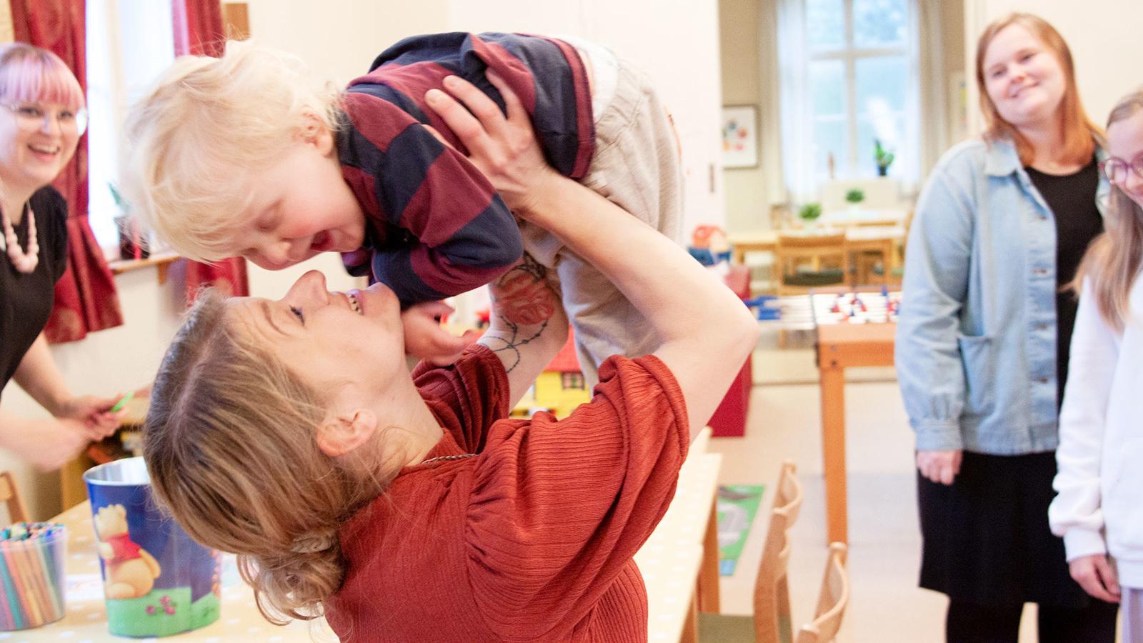 En mamma hissar skrattande sin leende lille son upp i luften inne på Frälsningsarmén i Eksjö. Runt omkring dem står Pernilla och Lina och tittar leende på.