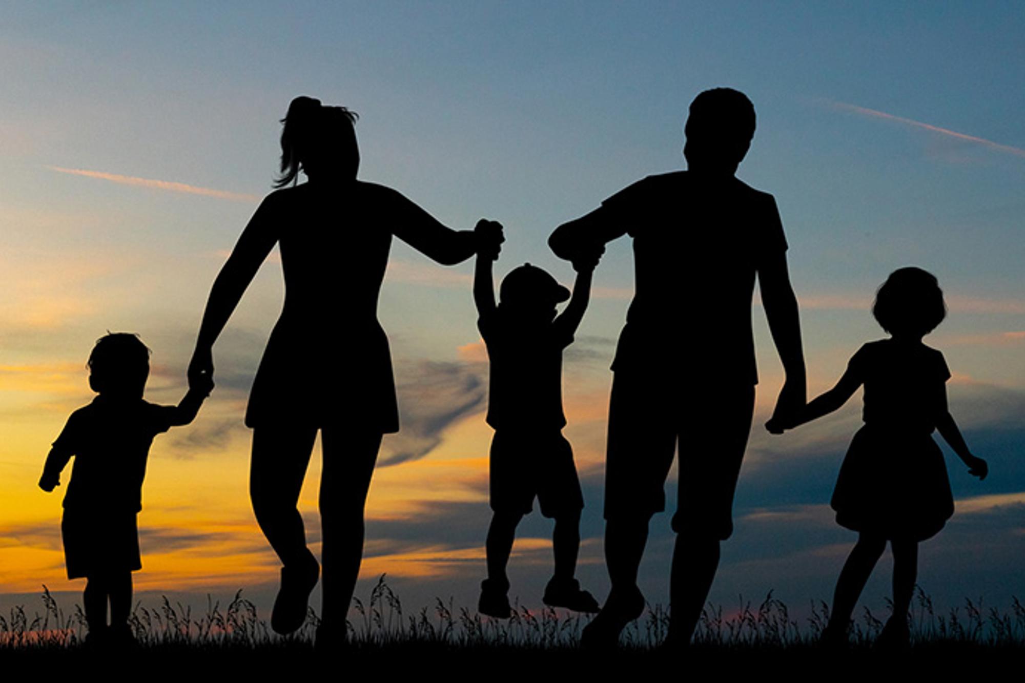 Siluetten mot en himmel med solnedgång av en kvinna och en man som tillsammans håller tre barn i händerna.