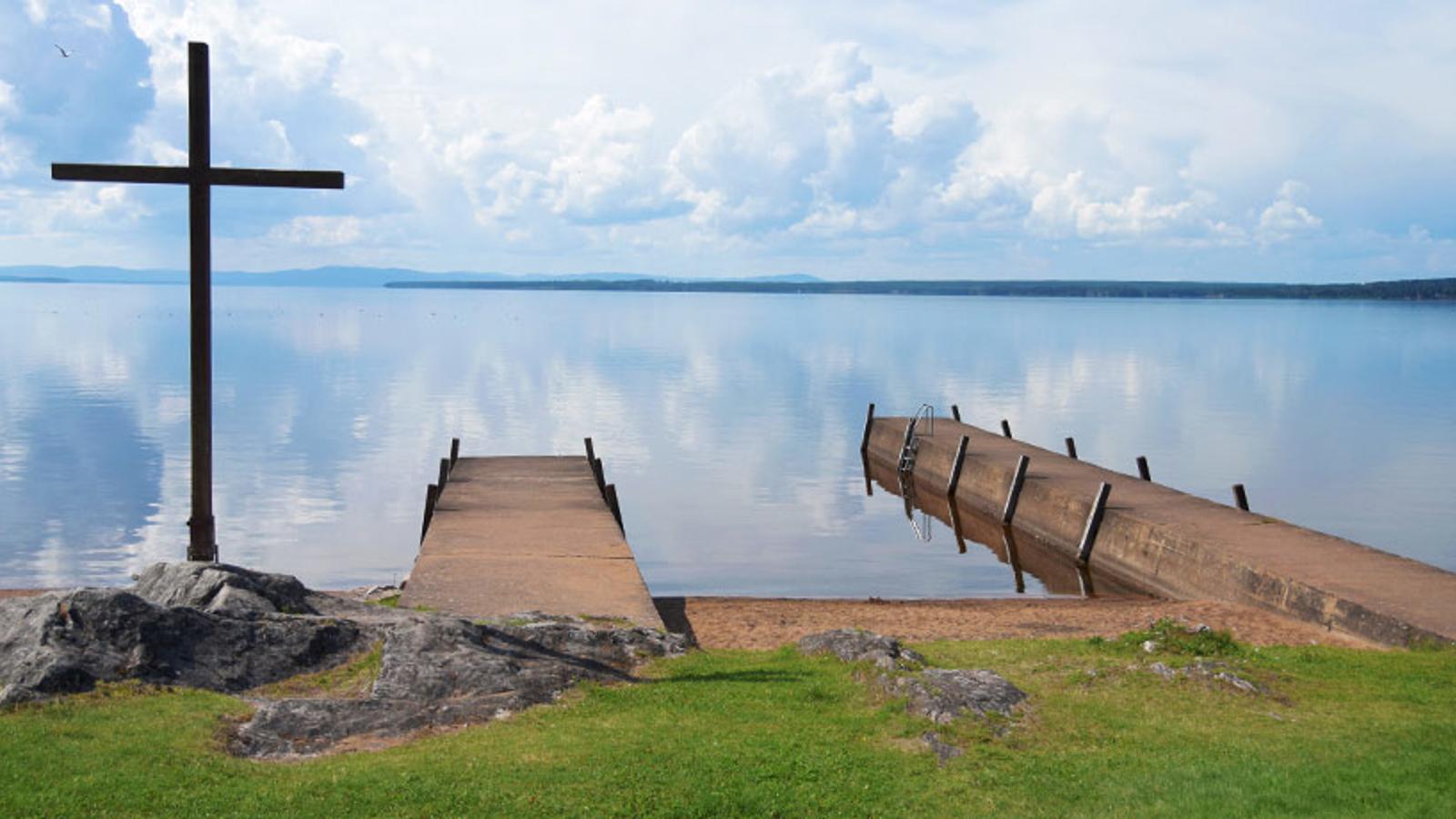 Klippor/stenar i förgrunden samt ett stort kors och två bryggor vid strandkanten på sjön Siljan som syns i bakgrunden.