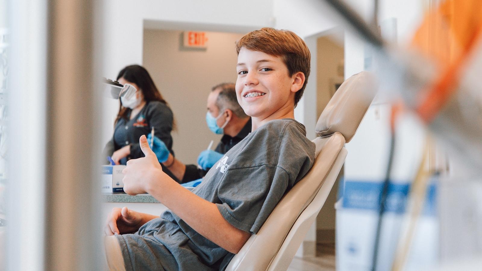 En pojke som sitter på en stol hos tandläkaren och gör tummen upp med ena handen.